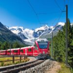 Un tren roșu circulă pe o linie ferată înconjurată de munți și păduri verzi.