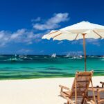 Plajă cu umbrele și șezlonguri, apă cristalină și bărci în depărtare.